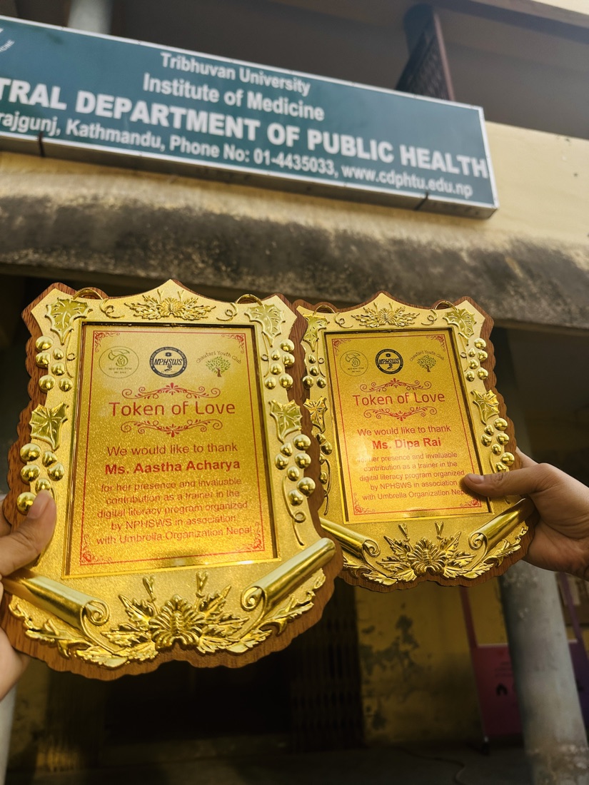 Two Token of Love appreciation plaques for Aastha Acharya and Dipa Rai held up in front of the CDPH sign